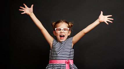 Happy little girl winning success. Cute kid standing with hands up and open mouth over black studio...