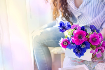 A woman is holding a gift with a bouquet of flowers in blue and pink. In a cozy house by the window. Happy woman. Postcard, free space for text.