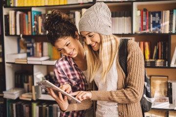 Pretty curly high school student girl checking tablet with a beautiful smiling female friend with a hat in the library.