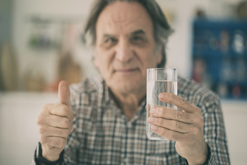 Senior man having glass of water and showing his thumb in kitchen