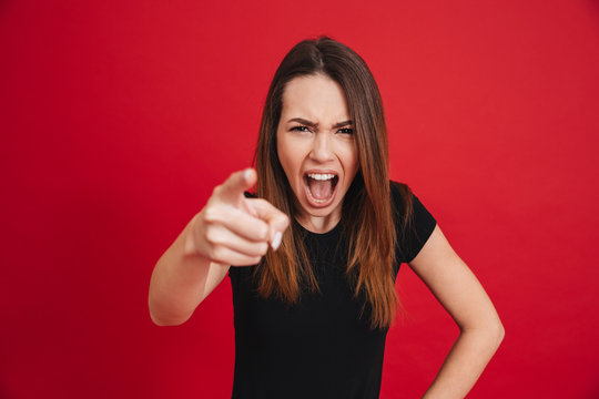 Portrait of emotional pretty woman in black t-shirt screaming and pointing index finger at camera like blaming you, isolated over red background