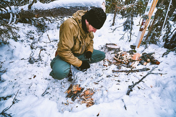 Young man in winter clothes is preparing a kindling for a campfire in winter in the tundra forest