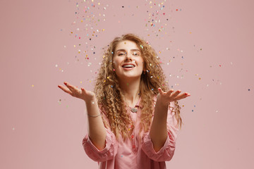 Young cheerful beautiful blonde girl in pink dress smiling with handful of confetti over pink...