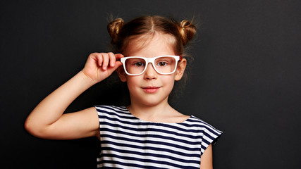 Portrait of beautiful kid putting on glasses on black background. Optometry and eye care concept. 