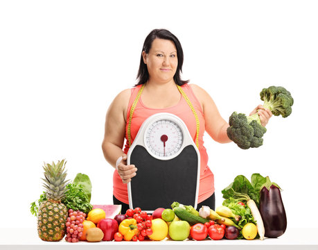 Overweight woman holding a weight scale and a broccoli dumbbell behind a table with fruit and vegetables