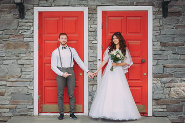 Stylish bride and groom posing on the background of the red door. Weddings in rustic style