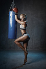 Portrait of sexy hot blonde boxer woman in red boxing gloves and grey sports bra and panties standing near blue punching bag without shoes