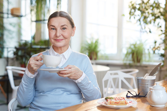 Smiling mature woman with cup of tea sitting by table and enjoying rest in cafe