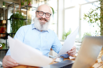 Cheerful mature man with business papers looking at camera with smile while working in cafe