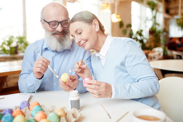 Aged couple with eggs and paintbrushes sitting by table and sharing creative ideas for Easter