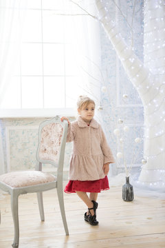 Adorable cute girl in red dress and pink coat is stayng near the chair in a fairy white room