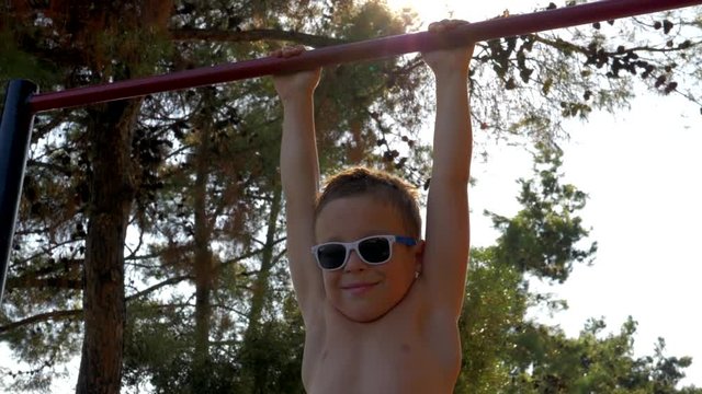 Slow motion shot of a smiling boy in sunglasses hanging on pull-up bar at outdoor sports ground
