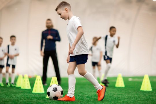 Little Football Player In Uniform Kicking Ball While Running Around Cone On The Field