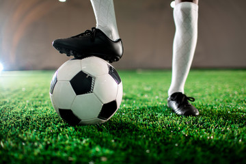 Young soccer player in boots and socks keeping one foot on ball while standing on green lawn