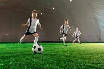 Foto op Plexiglas Three boys in uniform running after ball down green field while training indoors © pressmaster