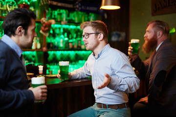 Friendly young man having talk while sitting by bar counter and drinking beer in pub