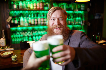 Excited bearded man with glass of foaming beer clinking with his friend in pub