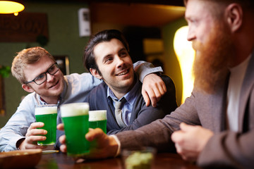 Two happy guys looking at bearded man during talk by glass of beer in pub