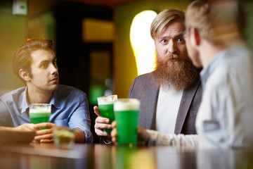 Three friendly men sitting by table or bar counter in pub and having beer