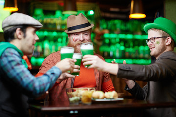 Friendly young men in hats sitting by table in pub and toasting with beer