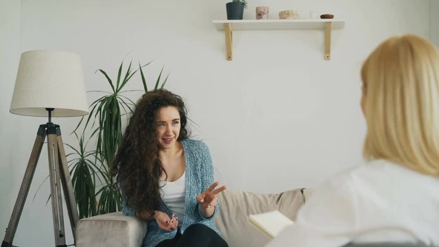 Smiling woman sitting on sofa and nervously talking to female psychoanalyst and gesturing hands in psychotherapist office indoors
