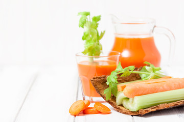 Carrot juice in beautiful glasses, cut carrot and green celery on wood bark bowl on white wooden background. Fresh vegetable drink. Close up photography. Selective focus. Horizontal banner
