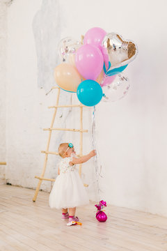 A Little Baby Girl In White Lavish Dress Is Standing In Living Room In The House Near The Decorative Staircase And Stretches To Bunch Of Brightly Colored Balloons. Concept Of Childrens Birthday Party