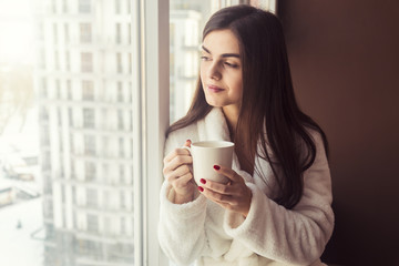 Beautiful girl wears white bathrobe holds cup of coffee sitting on the windowsill near window