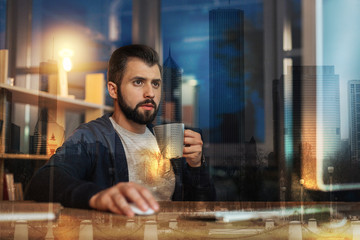 Evening activities. Calm attentive young man looking at the screen of a modern computer while sitting at the table with a cup in his hand