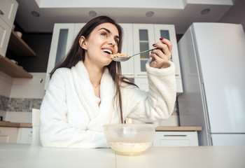 Fototapeta na wymiar Beautiful girl wears white bathrobe eating cornflakes with milk in the kitchen in the morning