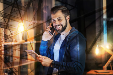 Good friend. Cheerful enthusiastic young man feeling happy while talking on the phone with his friend and holding a modern tablet in his hands