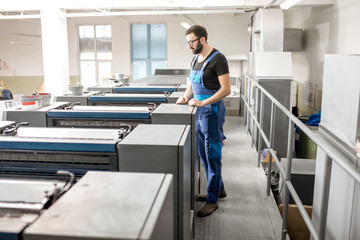 Worker in uniform supervising offset printing machine at the manufacturing