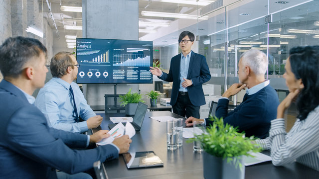 Asian Businessman Gives Report/ Presentation to His Business Colleagues, Pointing at the Results Showing Statistics, Pie Charts and Company's Growth On Wall TV Screen.