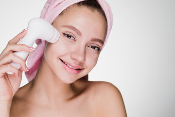 beautiful smiling girl with a pink towel on her head makes a deep cleansing of the face skin with a special electric brush