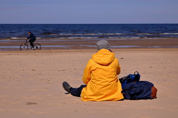 Samotna kobieta siedzi tyłem na piasku na plaży, w żółtej kurtce, obok niej leżą torby, w tle nieostry rowerzysta jedzie brzegiem morza, słonecznie - obrazy, fototapety, plakaty