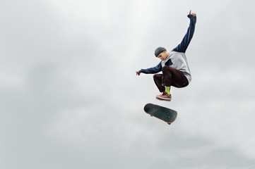 A skater teenager in a hat and a sweatshirt does a trick in the air with a flip of the board against a cloudy gray sky. The skater is isolated from other objects against the sky