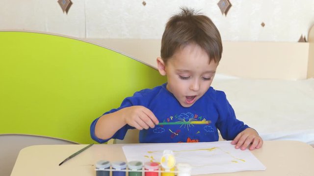 The child paints with watercolors on a white sheet of paper sitting at the table.