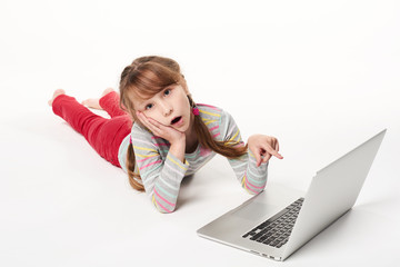 Surprised little girl lying on stomach on the floor with laptop, pointing at screen with amazement, over white background