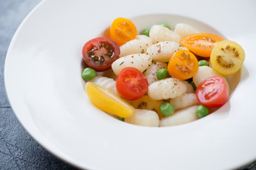 Closeup of boiled potato gnocchi served with fresh sliced tomatoes and green peas in a white plate, selective focus