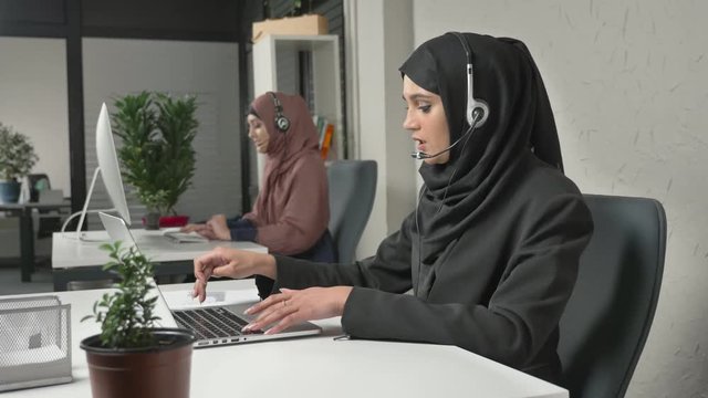 A young beautiful girl in a black hijab is talking on the headset, answering calls in call center. Arab women in the office. 60 fps