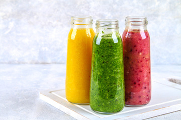 Green, yellow, purple smoothies in bottles of berries, greens, oranges on a white board on a gray table.