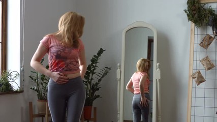 Charming middle aged redhead woman admiring her body shape in the mirror and slapping her buttocks in domestic interior. Cheerful woman looking at her reflection in the mirror with positive emotion.