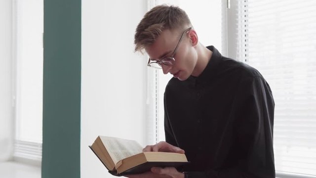 Young serious man is reading a bible against a window