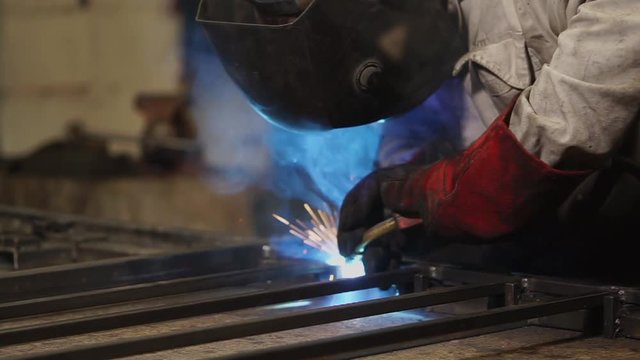 Welding operator is working in a forging manufactory. He is welding beams on a work table, close-up of his hands and mask