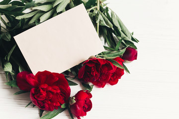 beautiful red peonies bouquet and craft paper card on white wooden rustic background. modern greeting mock up with space for text . top view. happy womens day or mothers