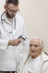 Lekarz w białym fartuchu mierzy temperaturę termometrem laserowym bardzo starej siwej kobiecie. Brodaty lekarz w białym kitlu ze stetoskopem na szyi mierzy temperaturę bardzo starej siwej kobiecie ter
