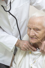 The internist doctor in a white dressing gown examines the lungs of a very old gray woman with a stethoscope. An internist doctor examines the lungs of a very old gray-haired woman sitting in a chair 