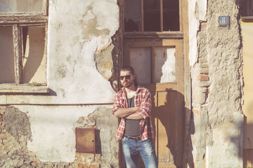 Man with beard and long hair posing in front of a ruined house.