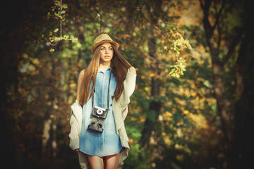 Modern young female photographer in autumn in park. Gorgeous girl in denim dress with vintage film camera outside in nature on sunny day.