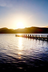 very long wooden jetty, jutting out into a calm blue wooden lake with mountains sunsetting in background